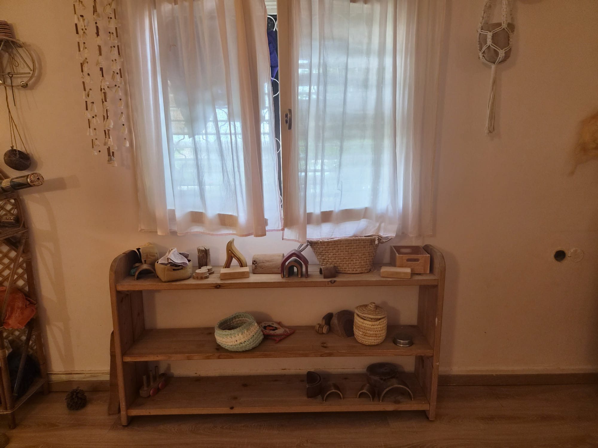 מדף צעצועי עץ טבעיים מתחת לחלון עם וילון לבן